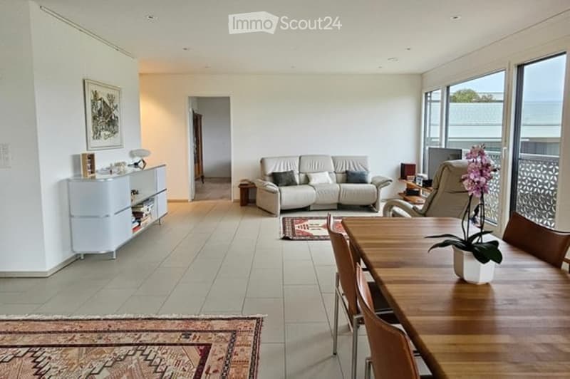 Bright, open living and dining area with large windows and a balcony view. Features a white sofa, wooden dining table with chairs, a cabinet, and decorative elements.