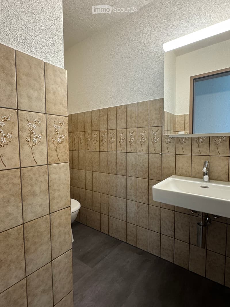 A small bathroom with white tile, tiled walls with floral patterns, a toilet, a white sink with a faucet, and a mirror.