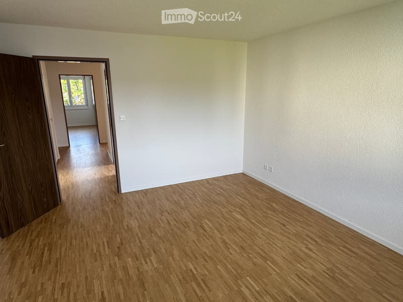 An empty room with white walls and wooden floor. It has an open door leading to another room with a window. The room has light switches and outlets on the wall. There is a hallway behind the open door, leading to a room with another window.