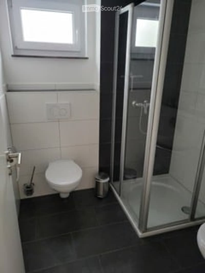 Modern bathroom with a toilet, glass-enclosed shower, and a small trash can. The walls are tiled in white and black, and the floor is dark gray.