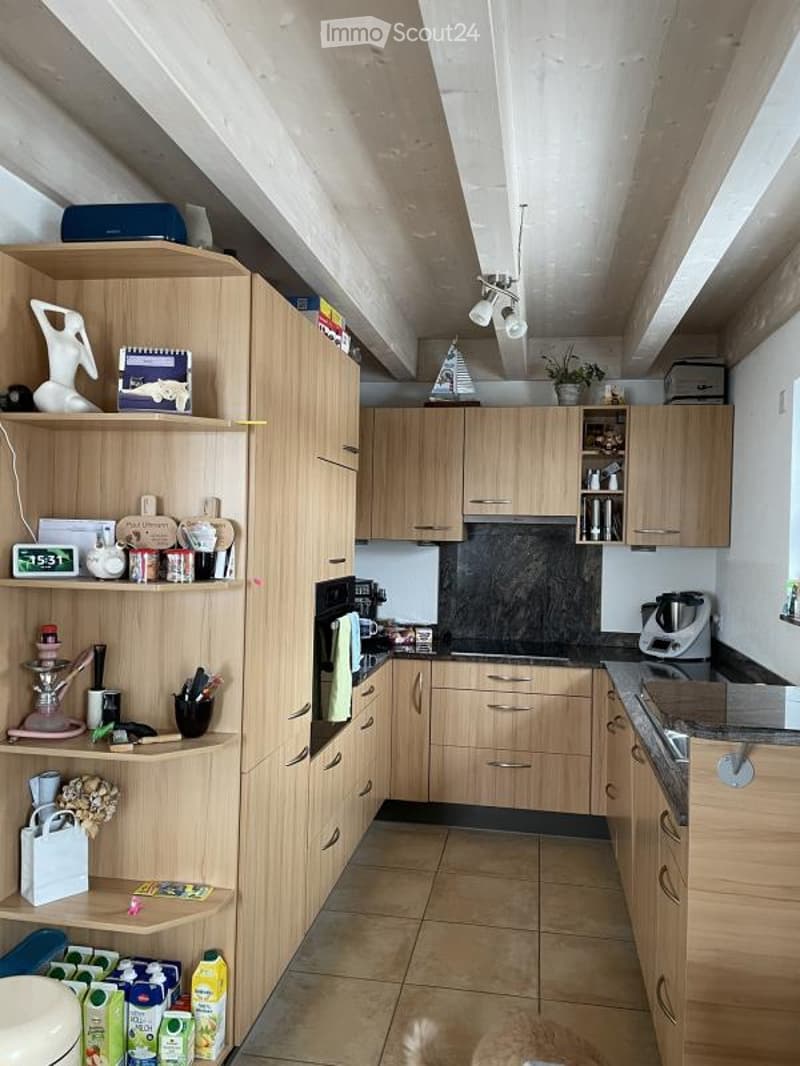 Modern kitchen with light wooden cabinets, stainless steel appliances, and a tiled floor. Shelves hold various items including a blender, cups, and decorative objects.