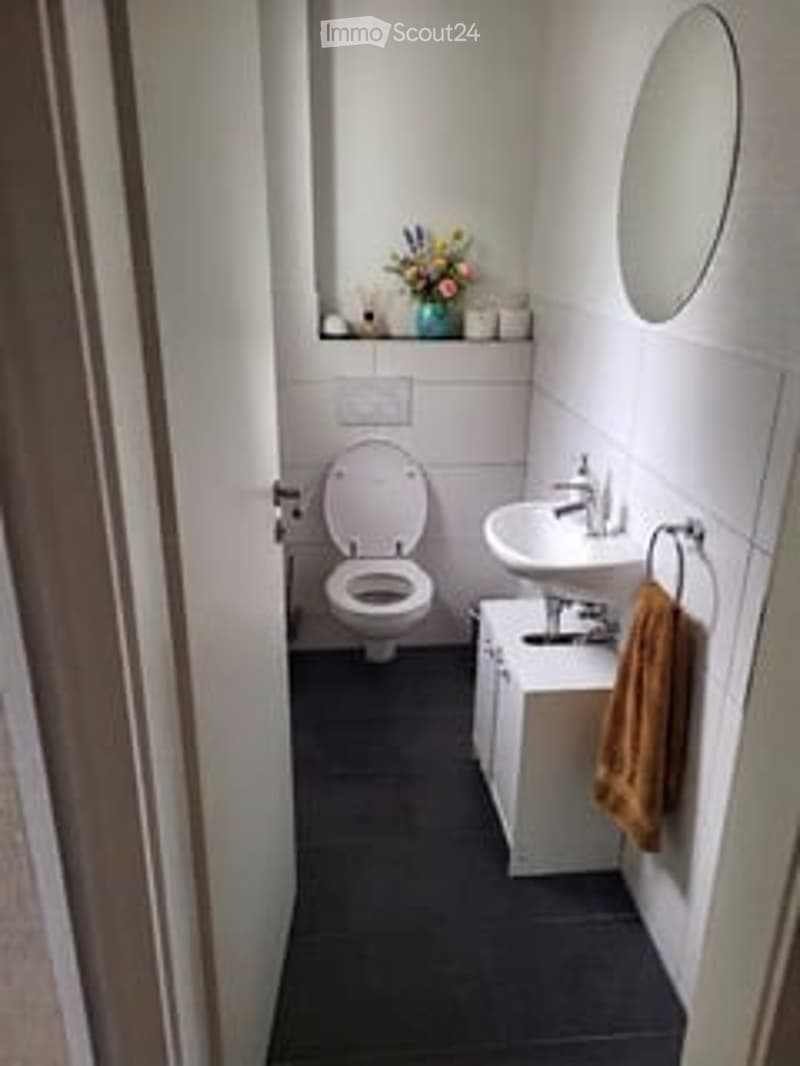 Modern bathroom with white tiles, black floor, toilet, sink, mirror, and a small cabinet with a towel hanging on the side.