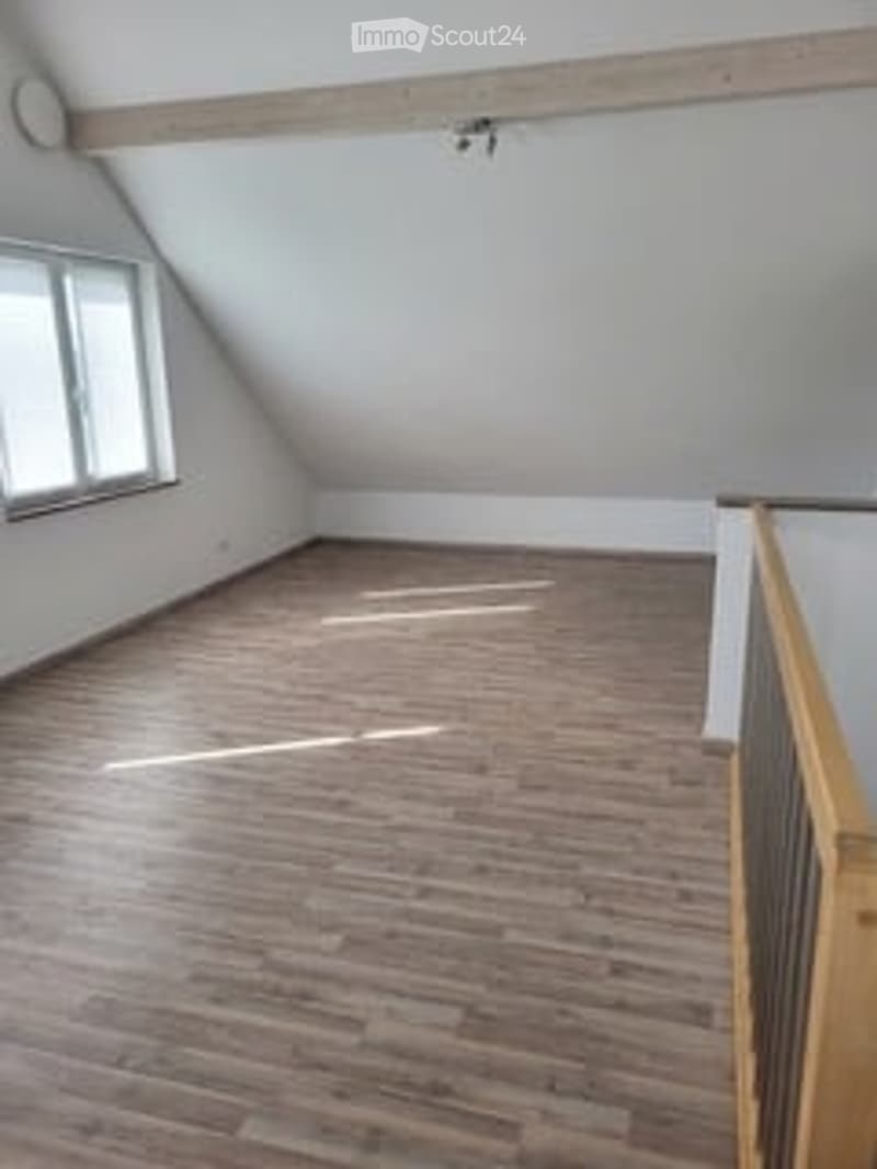 Empty attic room with wooden flooring, white walls, and large windows allowing natural light. No furniture or equipment present.