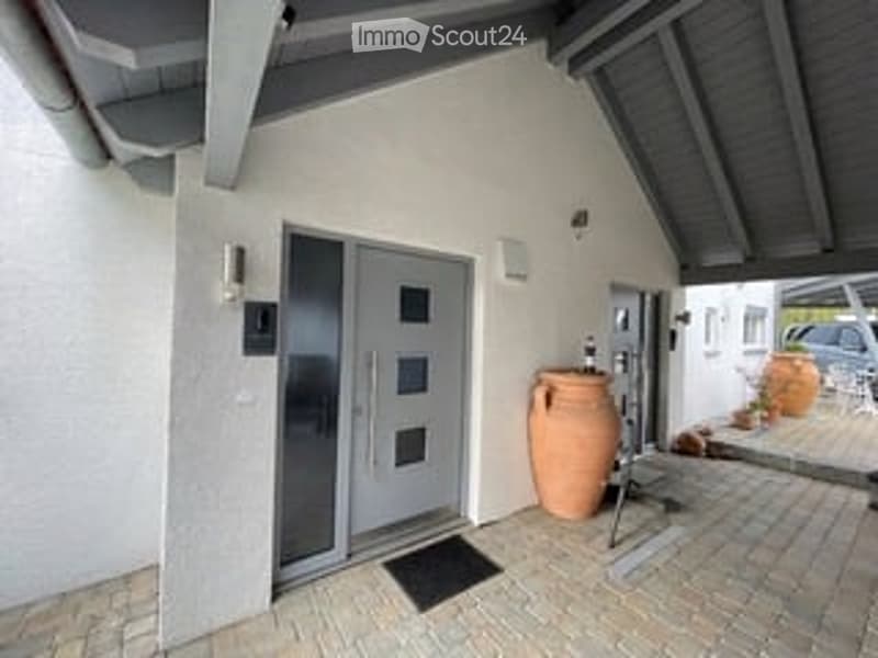 Front view of a modern house with a grey front door, white walls, and a small covered entrance area. A large terracotta vase is placed near the door, and a paved brick walkway leads to the entrance.
