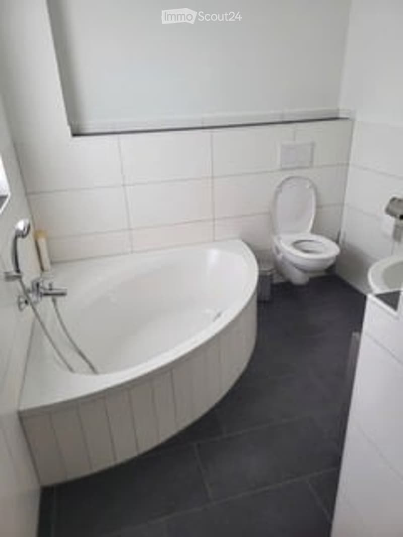 Modern bathroom with a white bathtub, toilet, and sink. The walls are tiled in white, and the floor is dark grey.