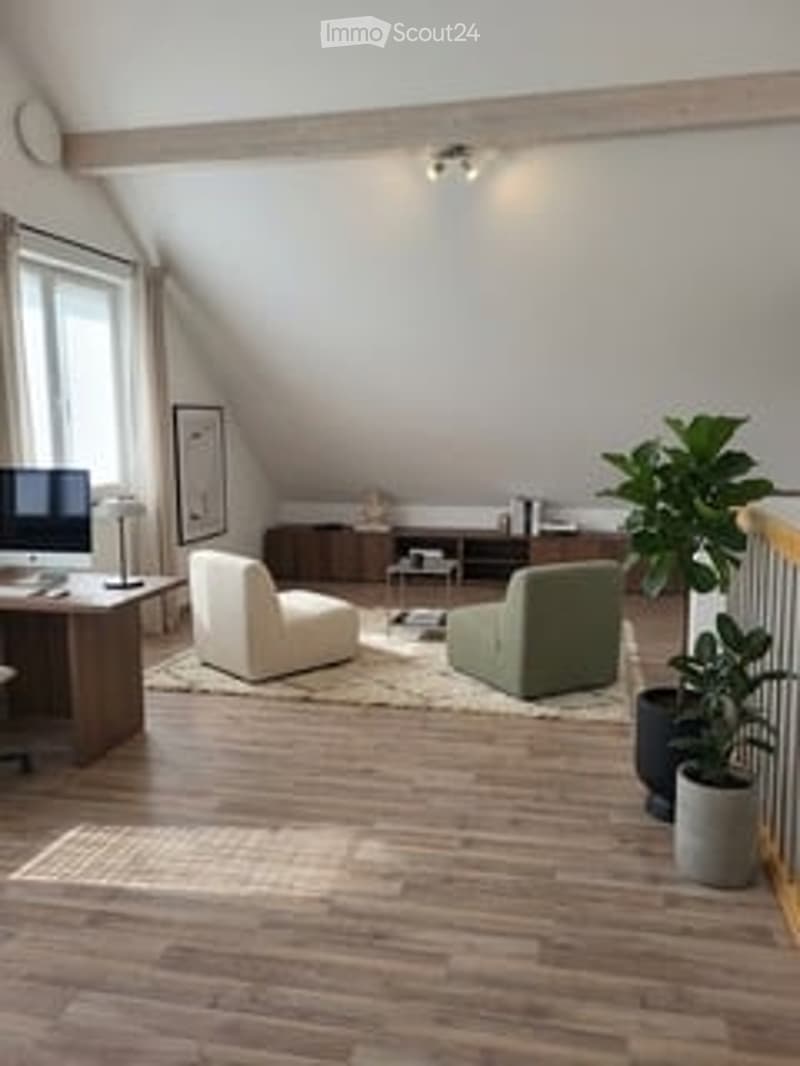 Bright living room with wooden flooring, large windows, a white armchair, green sofa, a small table, and a potted plant. A computer screen is visible on a desk in the corner.