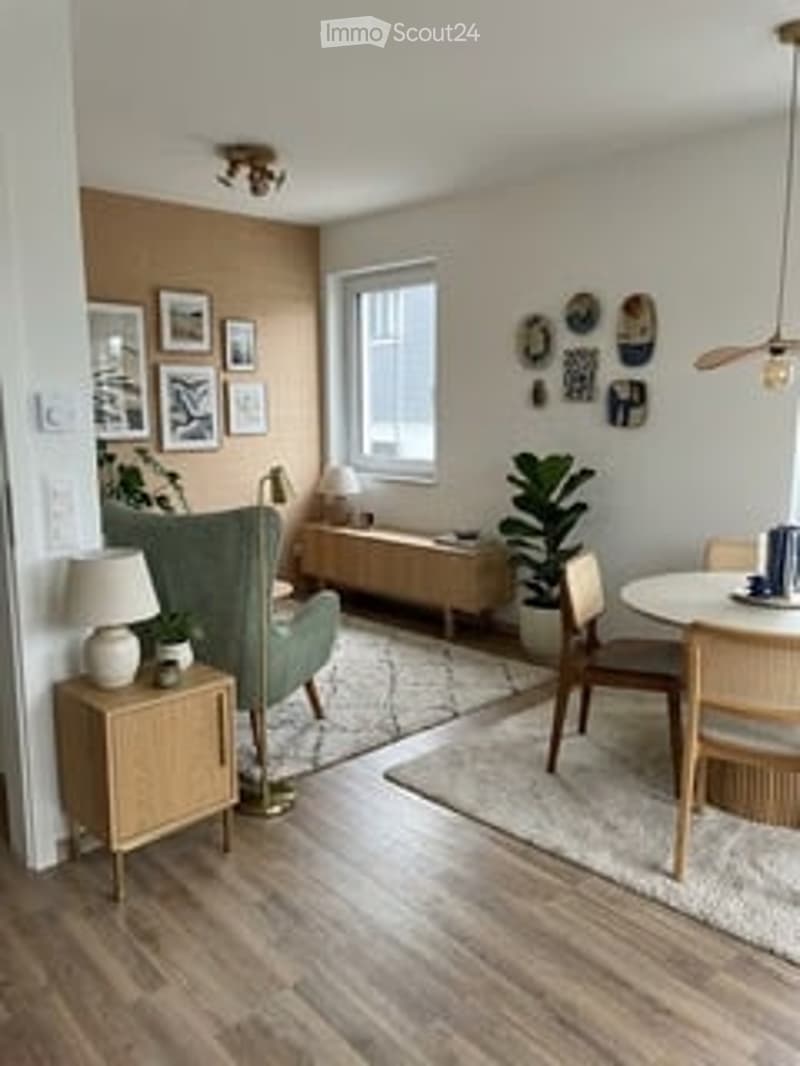 Bright, modern living and dining area with wooden flooring, large window, green sofa, coffee table, side table, dining table with chairs, pendant light, and decorative wall art.