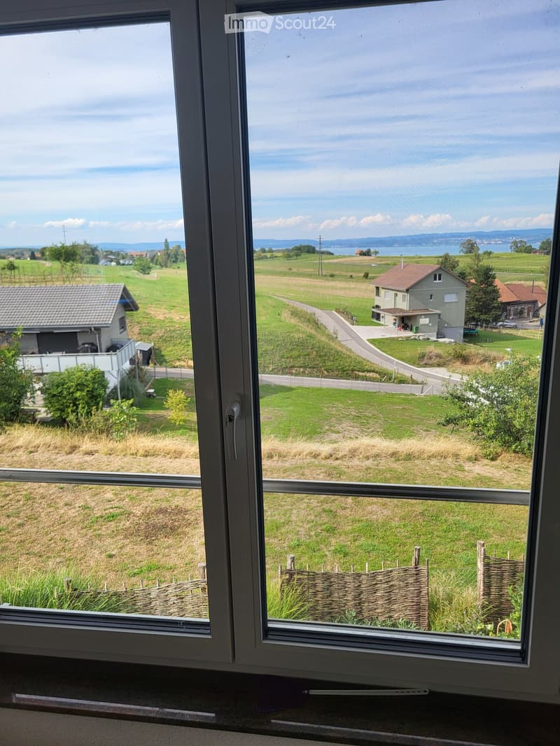 View from a window showing a rural landscape with fields, houses, and a distant body of water.