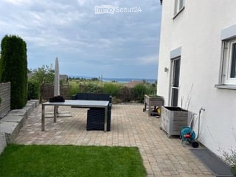 A modern patio area with a table, bench, umbrella, and storage box, surrounded by a neatly trimmed lawn and a white building.