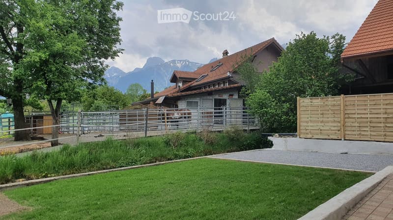 A well-maintained garden with a neatly trimmed lawn, a gravel pathway, and a wooden fence. A stable with a horse is visible in the background, along with a large house and a scenic mountain view.