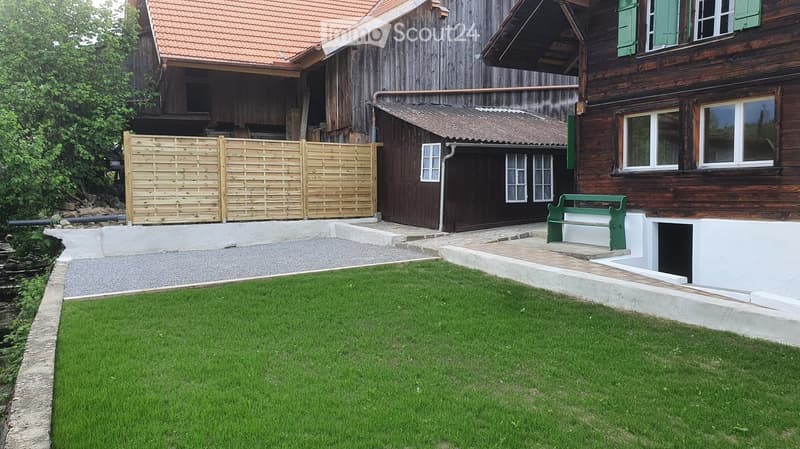 Well-maintained garden with green lawn, wooden fence, and traditional wooden house with green shutters and red-tiled roof.