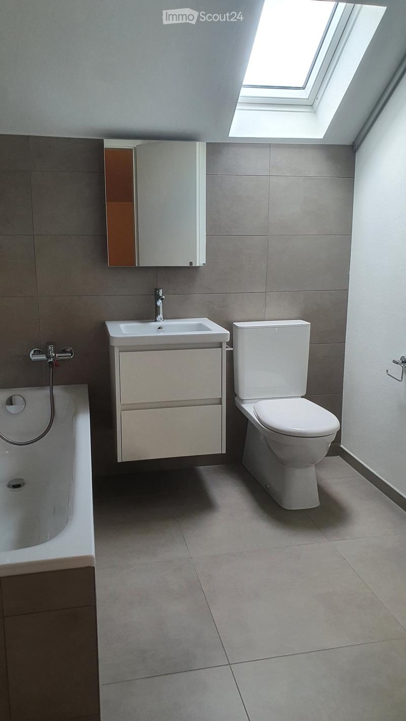 Modern bathroom with a bathtub, toilet, sink, and mirror. The room features grey tiles and a skylight window at the top.