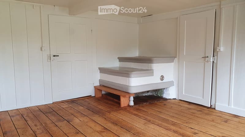 Empty room with wooden flooring, white walls, and three doors. A small wooden bench with a mattress is positioned against one wall.