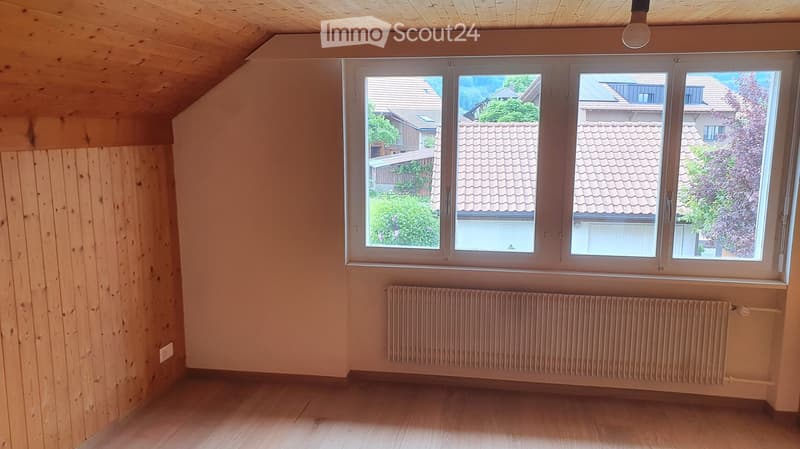 Empty room with wooden walls and floor, large window with view of neighboring houses and greenery, radiator below the window.