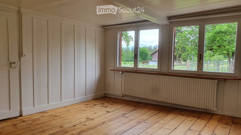 Empty room with wooden flooring, white paneled walls, large windows with a view of greenery, and a radiator below the windows.