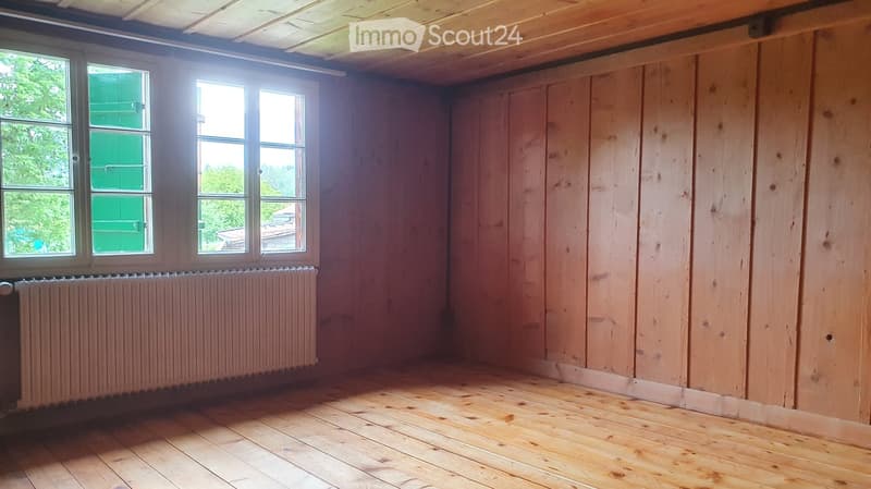 Empty room with wooden walls and floor, large windows, radiator, and exposed wooden ceiling beams.
