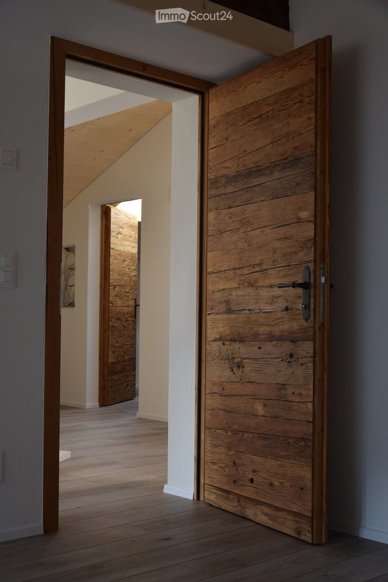 A modern hallway with wooden flooring, white walls, and a large wooden door partially open, revealing a glimpse of another room with exposed wooden beams and a slanted ceiling.