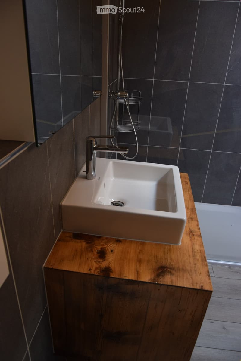 Modern bathroom with a white sink on a wooden vanity, chrome faucet, and a glass shower enclosure with dark tiles.