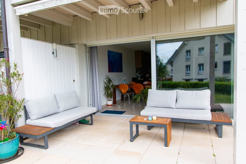 A modern outdoor patio with a white sofa, wooden coffee table, and chairs, connected to an indoor dining area with a table and chairs.