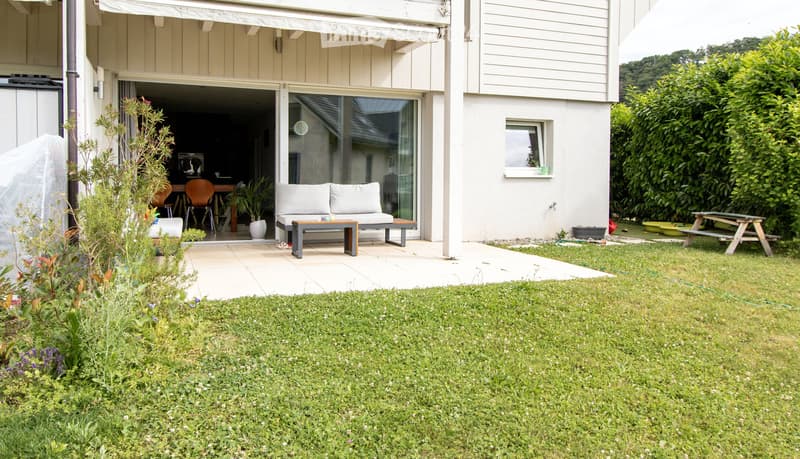 Garden area with a well-maintained lawn, a patio with outdoor seating, and a small wooden picnic table. The house features large glass doors leading to the patio, and there are various plants and shrubs around the area.