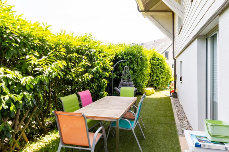 A small, enclosed patio area with artificial grass, a wooden table, colorful chairs, and a hanging egg chair. The space is surrounded by lush green hedges and has a gravel border along the wall.