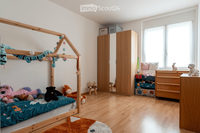 A well-lit children's bedroom with a wooden bed frame, colorful stuffed animals, a wardrobe, and a small table with toys.