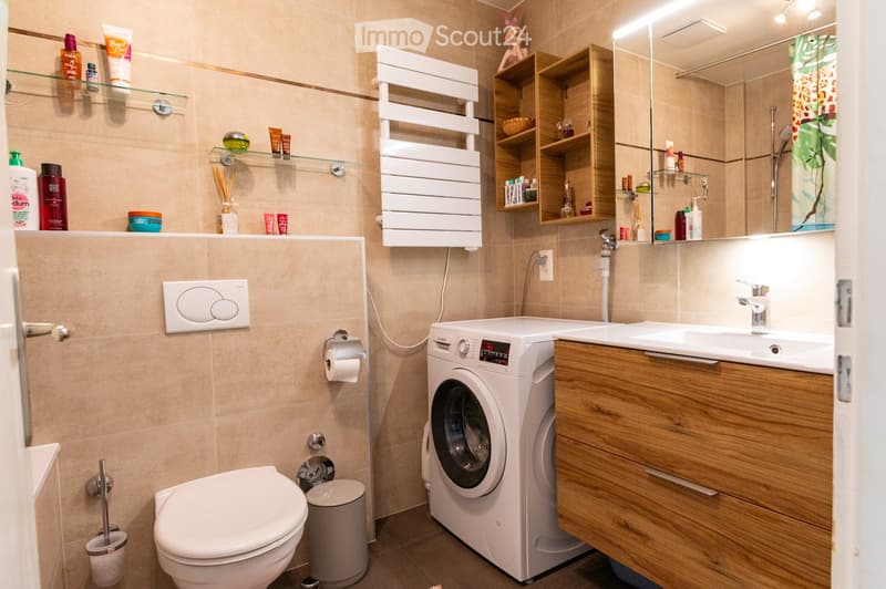Modern bathroom with beige tiles, a toilet, a sink with a wooden cabinet, a washing machine, shelves with toiletries, and a mirror above the sink.