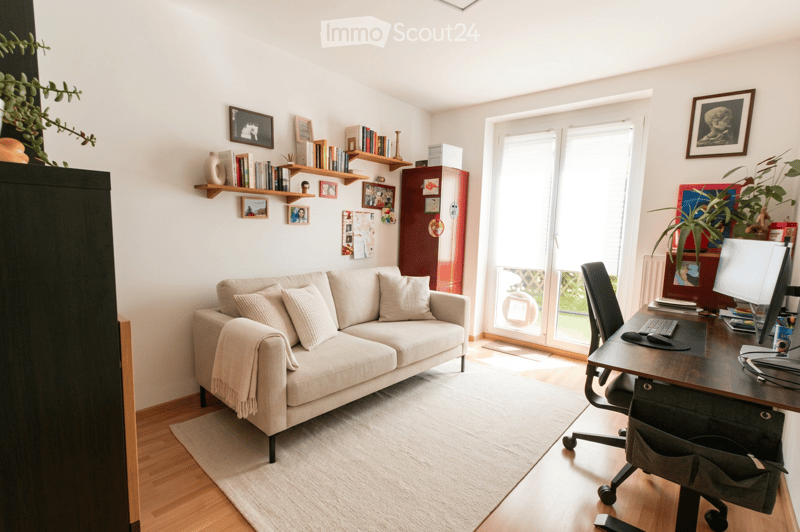 Bright living room with a beige sofa, a desk with a computer, and shelves filled with books and decorative items. Large windows and a sliding glass door lead to a balcony.