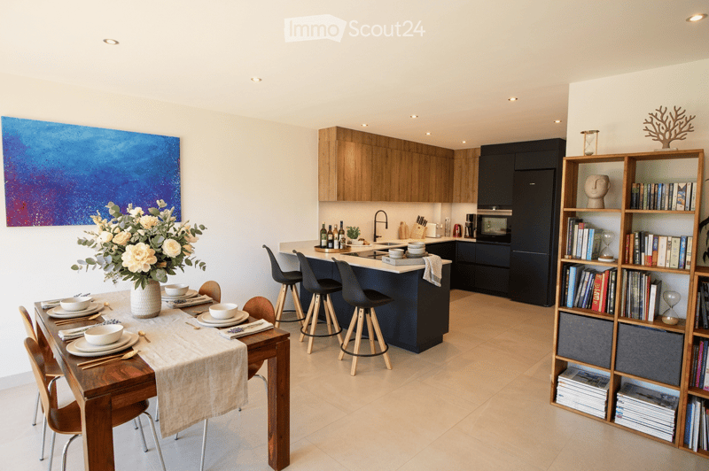 Modern kitchen and dining area with wooden cabinets, a central island, dining table with chairs, and a bookshelf filled with books and decorative items.