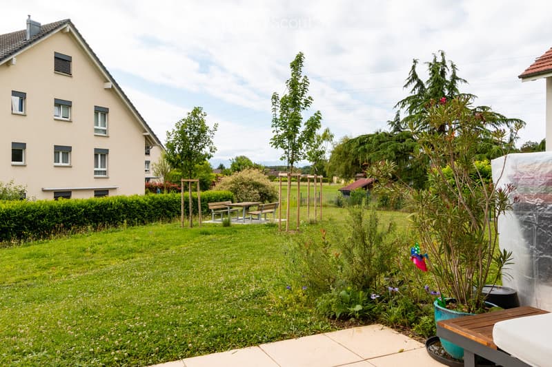 Well-maintained garden with green lawn, various plants, a small seating area with a table and benches, and a wooden fence. A single-story house with multiple windows is visible in the background.