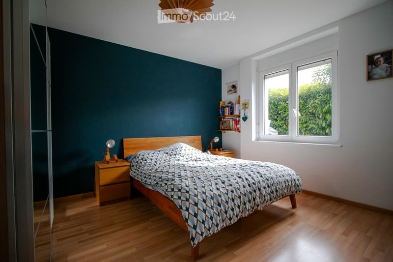 Modern bedroom with wooden flooring, dark green accent wall, large window with white frame, bed with patterned duvet, nightstand with lamp, bookshelf with books and decorative items.
