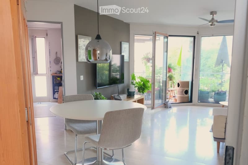 Modern dining area with a white round table, four grey chairs, a hanging pendant light, and a large window opening to a balcony with plants and a cat tree.