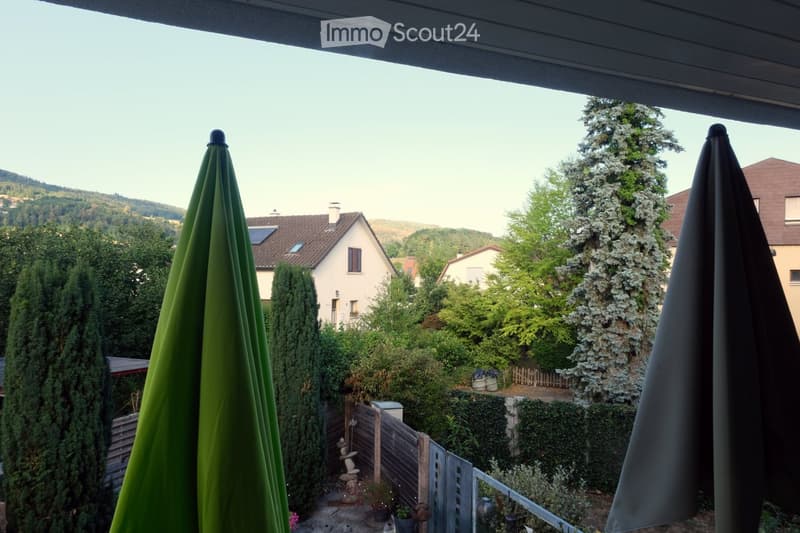 Balcony overlooking a landscaped garden with trees, a wooden fence, and neighboring houses in the background.