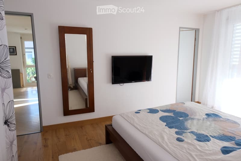 Modern bedroom with wooden flooring, a large bed with a blue and white floral duvet, a wall-mounted TV, a large mirror, and a small rug.