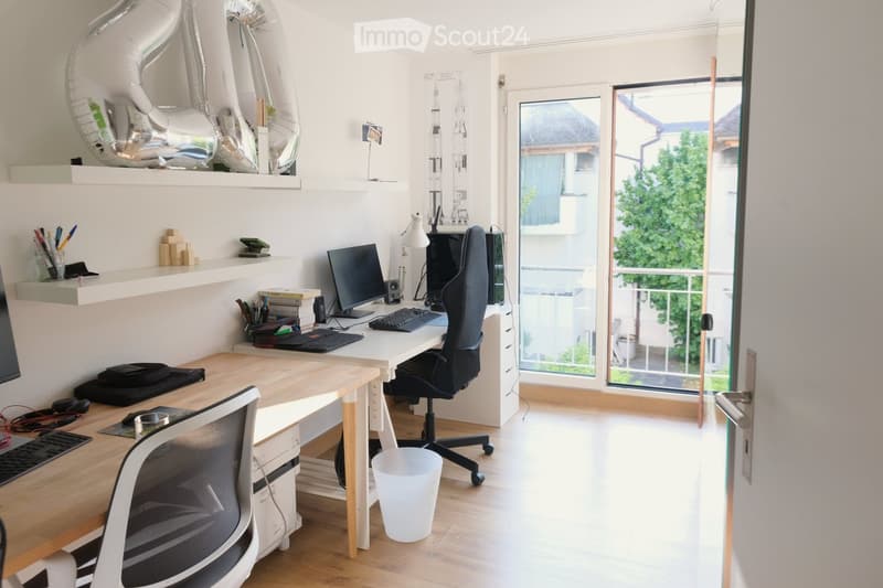 Modern, well-lit room with a desk, two chairs, computer setup, shelves with decorative items, and a large window with a view of greenery.