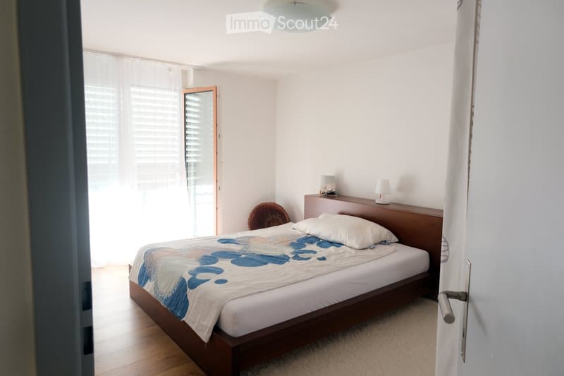 A modern bedroom with a wooden bed, white bedding, a nightstand with lamps, and a wicker chair near the window.