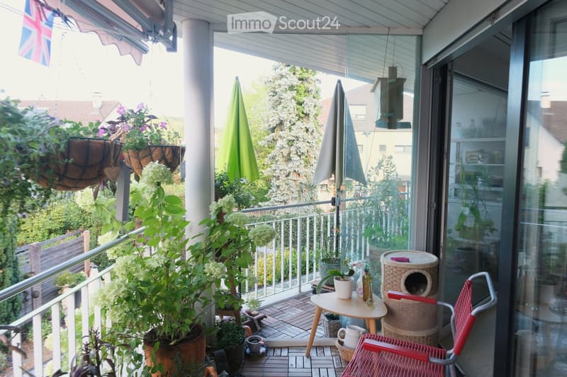 Balcony with wooden decking, potted plants, a small table with a chair, a cat tree, and two closed umbrellas.