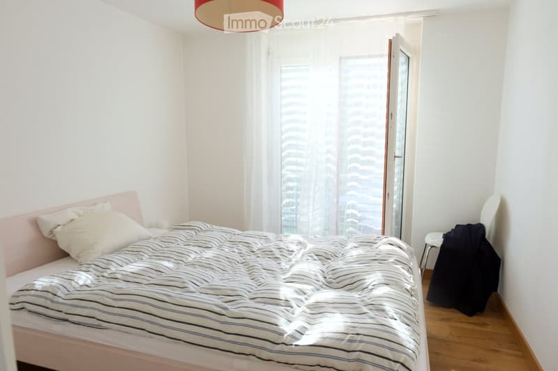 Bright bedroom with a white bed, striped duvet, pink headboard, and a chair with a jacket draped over it. Large windows with sheer curtains offer a view of city buildings.