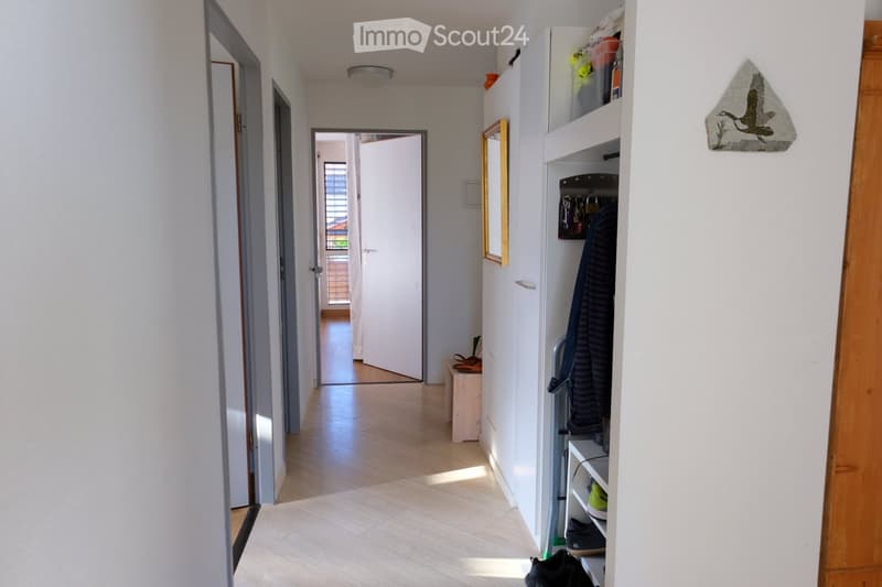 A clean, well-lit hallway with light wooden flooring, white walls, and a built-in storage unit holding shoes and other items. A mirror and a decorative wall piece are visible on the right side.