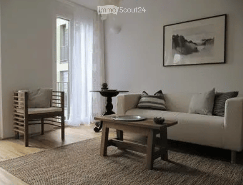 Modern living room with white walls, wooden floor, large windows, balcony access, sofa, coffee table, chair, small table, and picture frame.