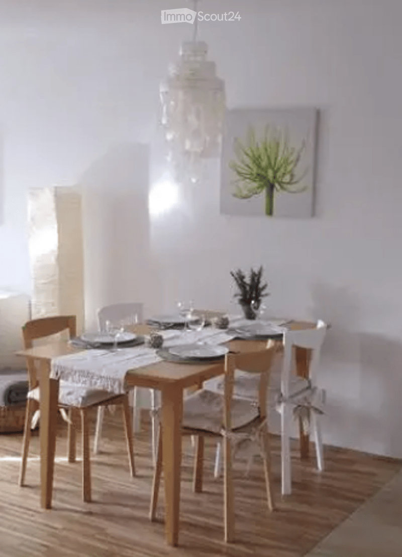 Dining room with a wooden dining table, six chairs, plates, cups, and a vase with flowers on the table. A chandelier hangs from the ceiling, and a picture frame is on the wall.