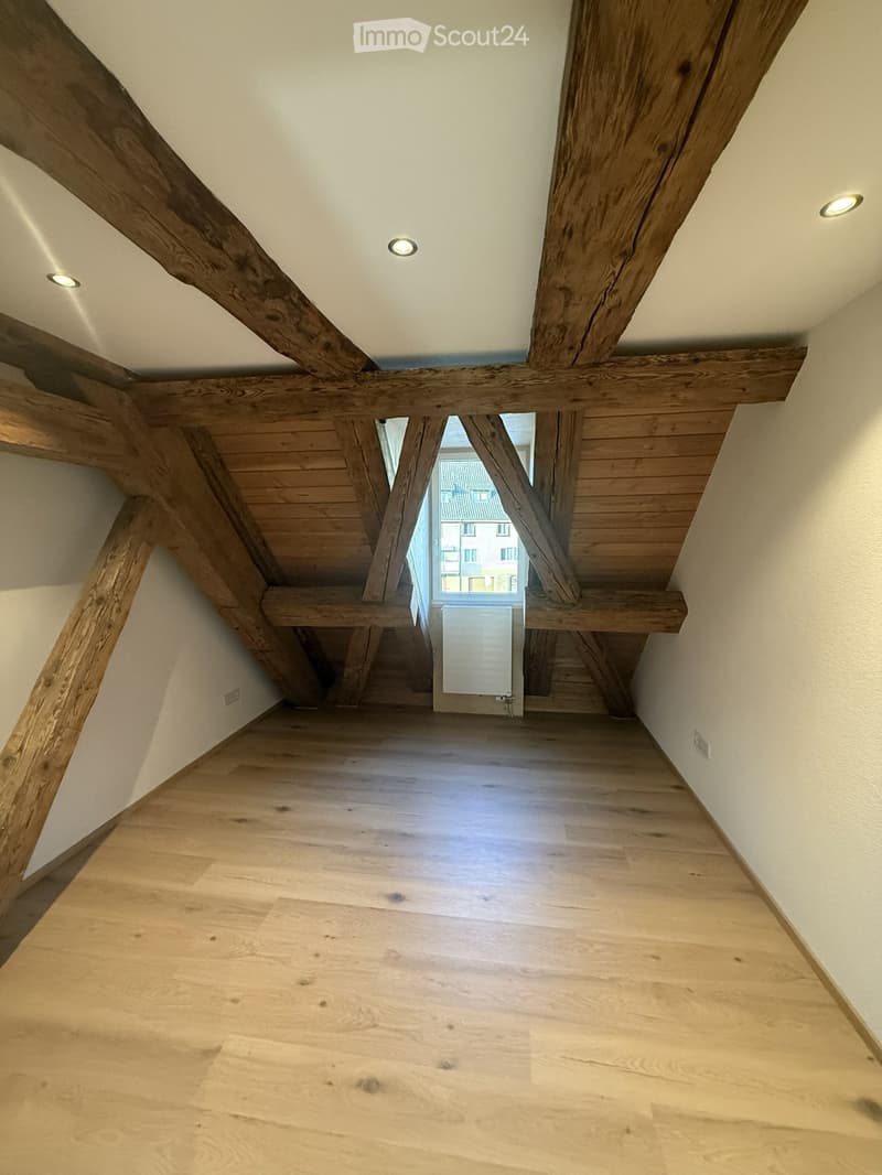 Empty hallway with wooden floor, exposed wooden beams, recessed ceiling lights, and a window at the end showing a view of the outside.