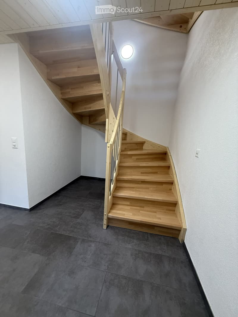 Wooden staircase with a modern railing, grey tiled floor, white walls, and a circular light fixture on the wall above the stairs.