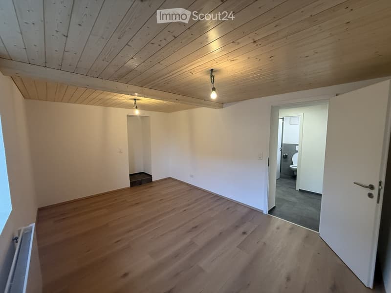 Empty room with wooden flooring, white walls, and exposed wooden ceiling beams. A doorway leads to a small bathroom with a toilet visible.