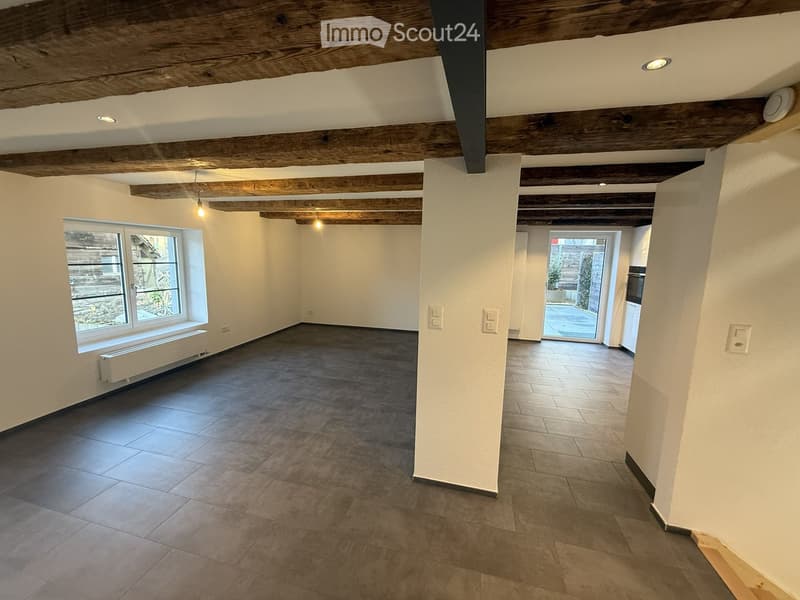 Empty room with exposed wooden beams on the ceiling, tiled floor, large window, and a doorway leading to another area.