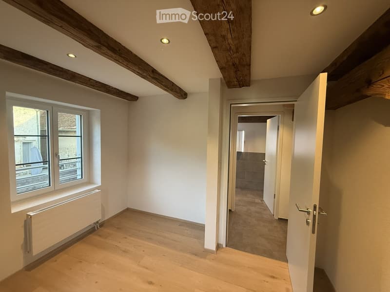 Empty room with wooden flooring, white walls, exposed wooden beams, and a window with a radiator below it. Open doors lead to another room.