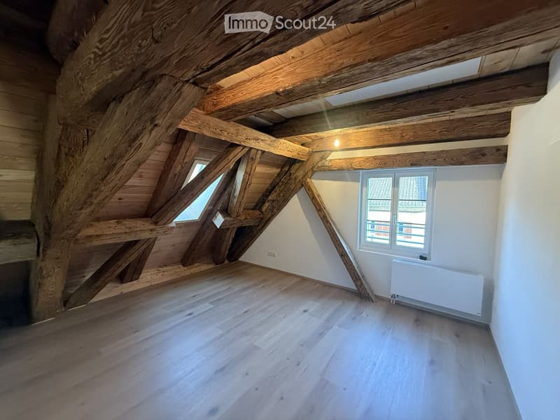 Empty attic space with wooden beams, wooden floor, white walls, and two windows, one small skylight and one larger window with a view of the roof.