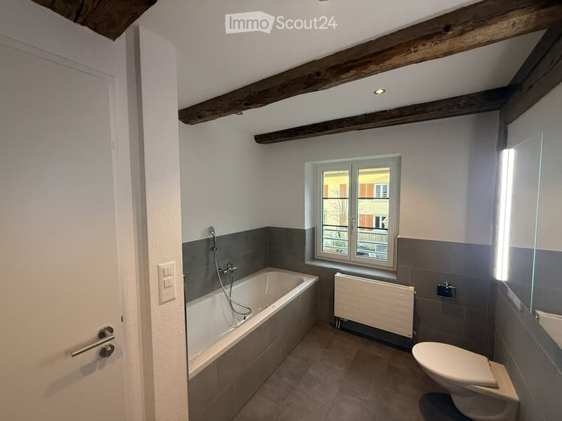 Bathroom with a bathtub, toilet, radiator, and a large window offering a view of the outside. Exposed wooden beams on the ceiling and modern grey tiles.