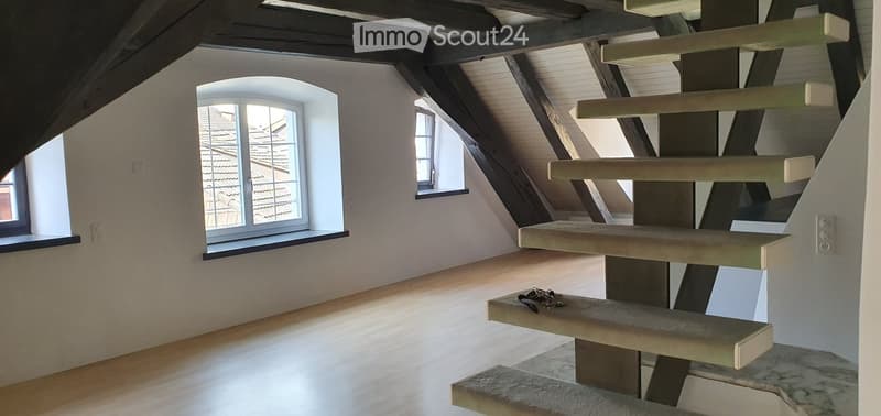 Empty attic space with wooden floors, exposed wooden beams, and large windows offering a view of the roof. A modern, minimalist staircase is visible.