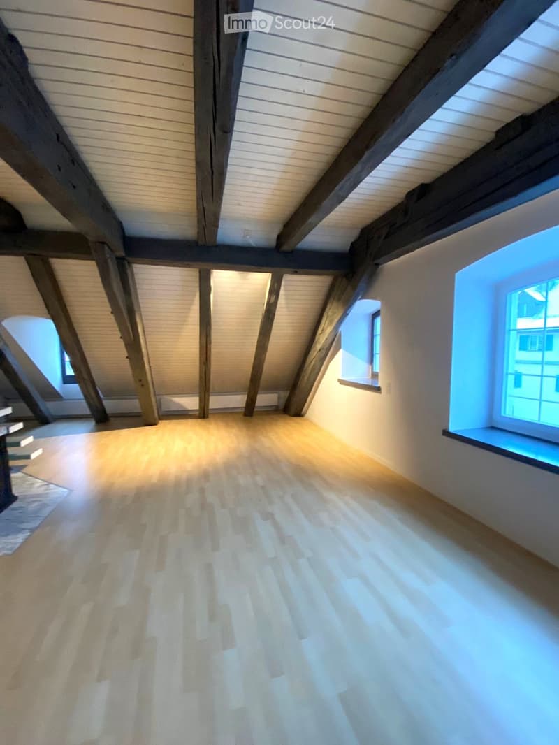 An empty attic space with wooden beams, exposed wooden ceiling, and wooden flooring. Three windows provide natural light.